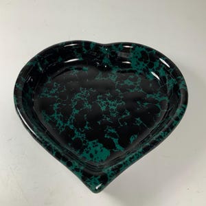 May include: Heart-shaped decorative dish with a glossy finish. The dish features a black base with a green, marbled pattern. The dish is empty and sits on a white surface. The dish is likely made of ceramic or a similar material.