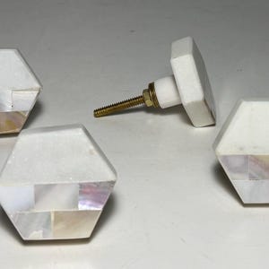 May include: Four hexagonal cabinet knobs featuring a smooth white top section and a lower section inlaid with iridescent mother-of-pearl tiles. Each knob includes a gold-toned metal screw post extending from the back for mounting purposes.