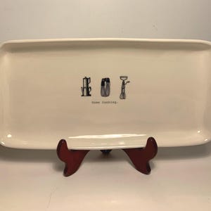 Puede incluir: Una fuente rectangular de cerámica blanquecina con esquinas redondeadas. Presenta dibujos lineales negros de una cafetera, una batidora y un batidor, con las palabras "Home Cooking" debajo. La fuente se exhibe sobre un soporte rojo oscuro.