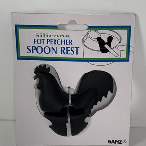 Op de afbeelding: Zwarte siliconen lepelhouder in de vorm van een haan, verpakt op een witte kaart. De kaart heeft de woorden "Silicone Pot Percher Spoon Rest" in groen en blauw. De lepelhouder is ontworpen om op de rand van een pot te rusten.