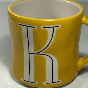 Puede incluir: Una taza de cer&aacute;mica amarilla con interior blanco y asa. La letra "K" se muestra en blanco con contornos negros en la parte delantera. La taza es de color amarillo brillante y s&oacute;lido. Adecuada para bebidas fr&iacute;as o calientes.