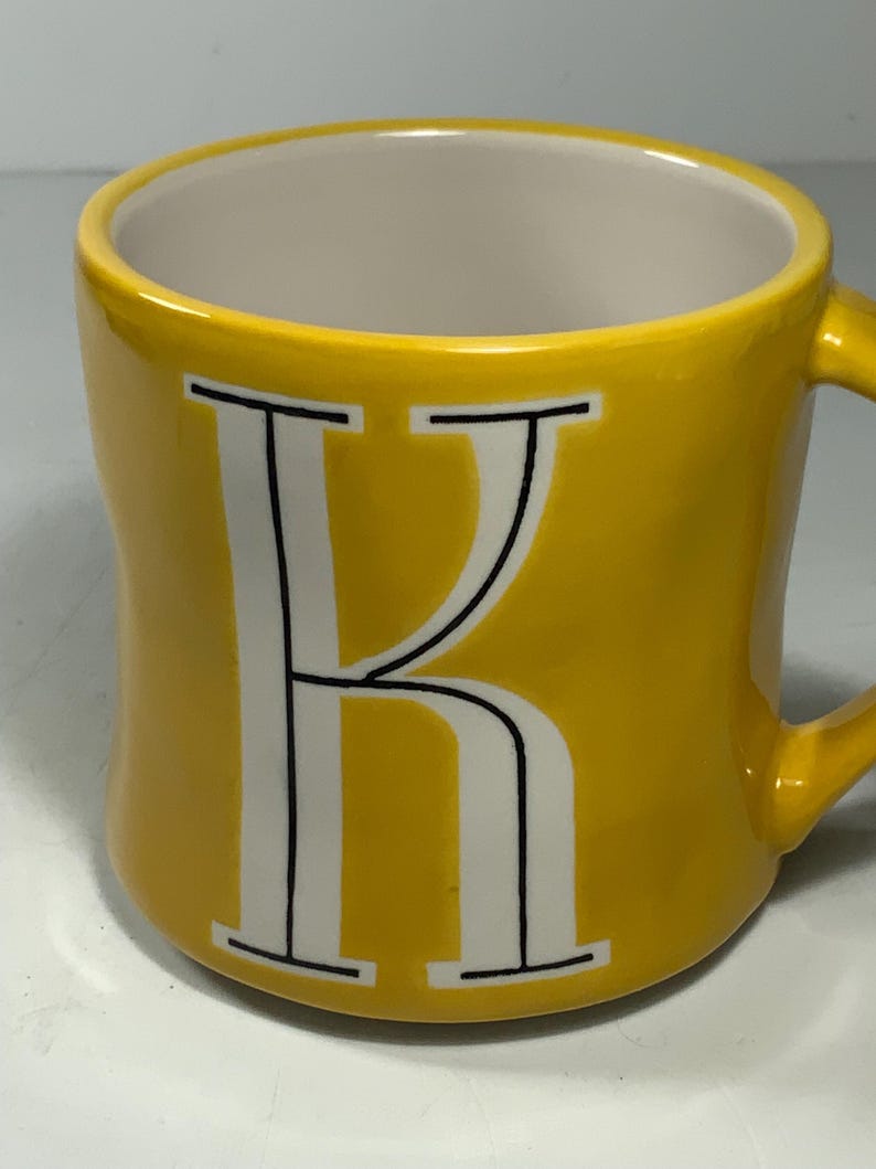 Puede incluir: Una taza de cer&aacute;mica amarilla con interior blanco y asa. La letra "K" se muestra en blanco con contornos negros en la parte delantera. La taza es de color amarillo brillante y s&oacute;lido. Adecuada para bebidas fr&iacute;as o calientes.