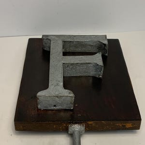 May include: A metal letter "F" mounted on a dark brown wooden plaque with a metal rod attached to the bottom. The letter is a dull silver color, and the wood has a rich, aged appearance. The piece is likely a decorative item.