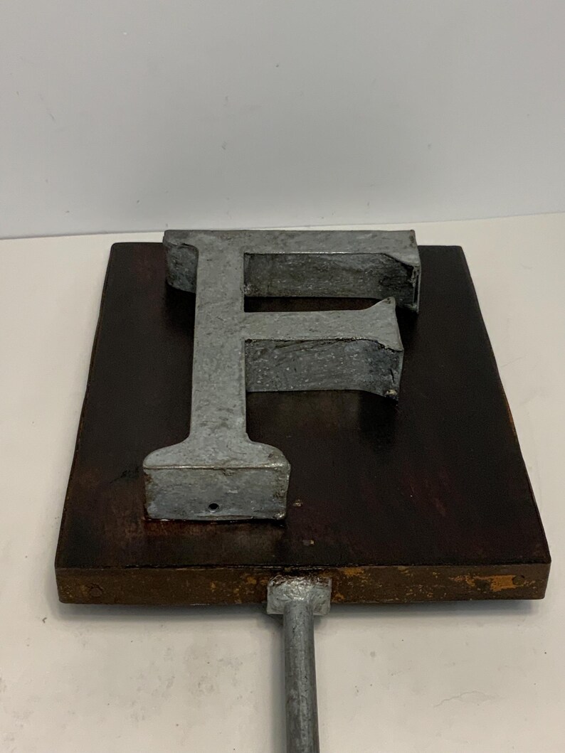 May include: A metal letter "F" mounted on a dark brown wooden plaque with a metal rod attached to the bottom. The letter is a dull silver color, and the wood has a rich, aged appearance. The piece is likely a decorative item.