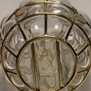 Op de afbeelding: Een heldere glazen vaas met een bolvorm, omhuld door een goudkleurige metalen kooi. Het glas heeft een bubbelachtige textuur en de metalen kooi heeft cirkelvormige en lineaire elementen. De vaas lijkt leeg te zijn.
