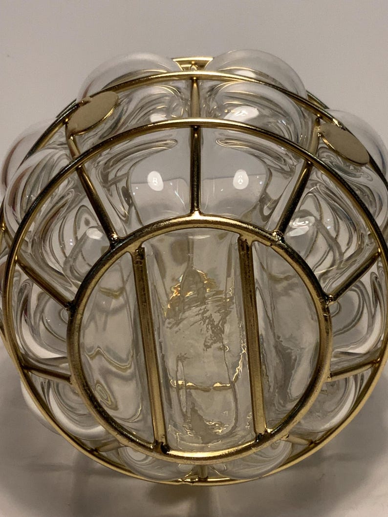 Op de afbeelding: Een heldere glazen vaas met een bolvorm, omhuld door een goudkleurige metalen kooi. Het glas heeft een bubbelachtige textuur en de metalen kooi heeft cirkelvormige en lineaire elementen. De vaas lijkt leeg te zijn.