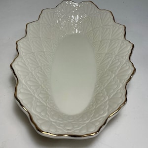 Puede incluir: Un plato ovalado de cerámica blanca con borde festoneado y acabado en oro. La superficie interior está decorada con un patrón repetitivo en relieve de hojas o pétalos que irradia desde el centro liso y plano hacia los bordes exteriores.