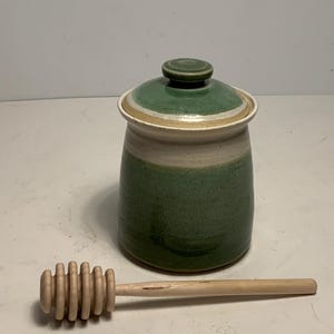 May include: A ceramic honey pot with a green and tan lid and body, and a wooden honey dipper. The pot is cylindrical with a rounded lid. The honey dipper is made of light-colored wood.