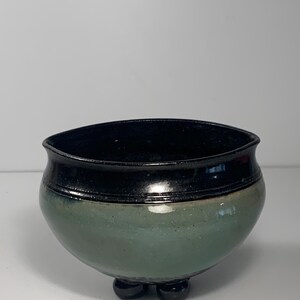 May include: A handcrafted ceramic bowl with a rounded body and three small feet. The bowl features a glossy black rim and a band of the same color, with a muted green-blue body. The interior is black.