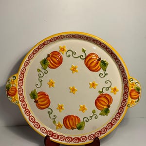 May include: Round ceramic serving platter with a cream centre, featuring orange pumpkins, green leaves, and yellow star-shaped flowers. The rim has a yellow border and a red floral pattern. Includes handles.