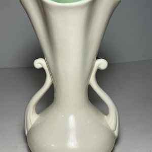 以下が含まれることがあります： クリーム色の陶器製花瓶で、広がった波状の縁と、両側に2つの対称的な曲線状のハンドルが付いています。花瓶の内側は淡いミントグリーンの釉薬で仕上げられ、外側は滑らかなオフホワイトの光沢のある質感になっています。