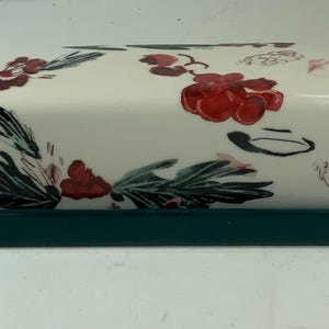 May include: A ceramic butter dish with a white lid featuring a floral design of red berries and green leaves. The base is a solid dark teal color, providing a contrasting frame for the decorative lid.