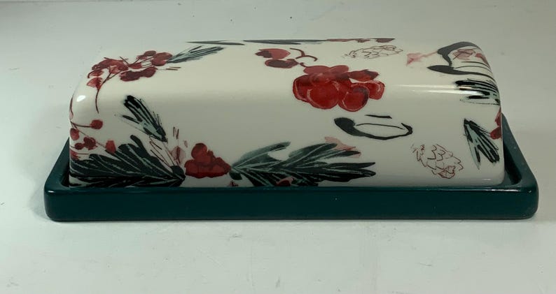 May include: A ceramic butter dish with a white lid featuring a floral design of red berries and green leaves. The base is a solid dark teal color, providing a contrasting frame for the decorative lid.