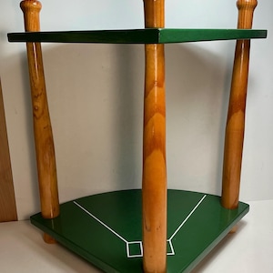 Pode incluir: Uma prateleira de canto de dois níveis com três tacos de beisebol de madeira verticais como pilares de suporte. As duas prateleiras triangulares são pintadas de verde escuro com linhas brancas formando um padrão de campo de beisebol.
