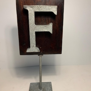 May include: A decorative wooden sign featuring the letter "F" in a silver-colored metal. The letter is mounted on a dark brown rectangular wooden plaque. The plaque is attached to a metal stand with a square base.