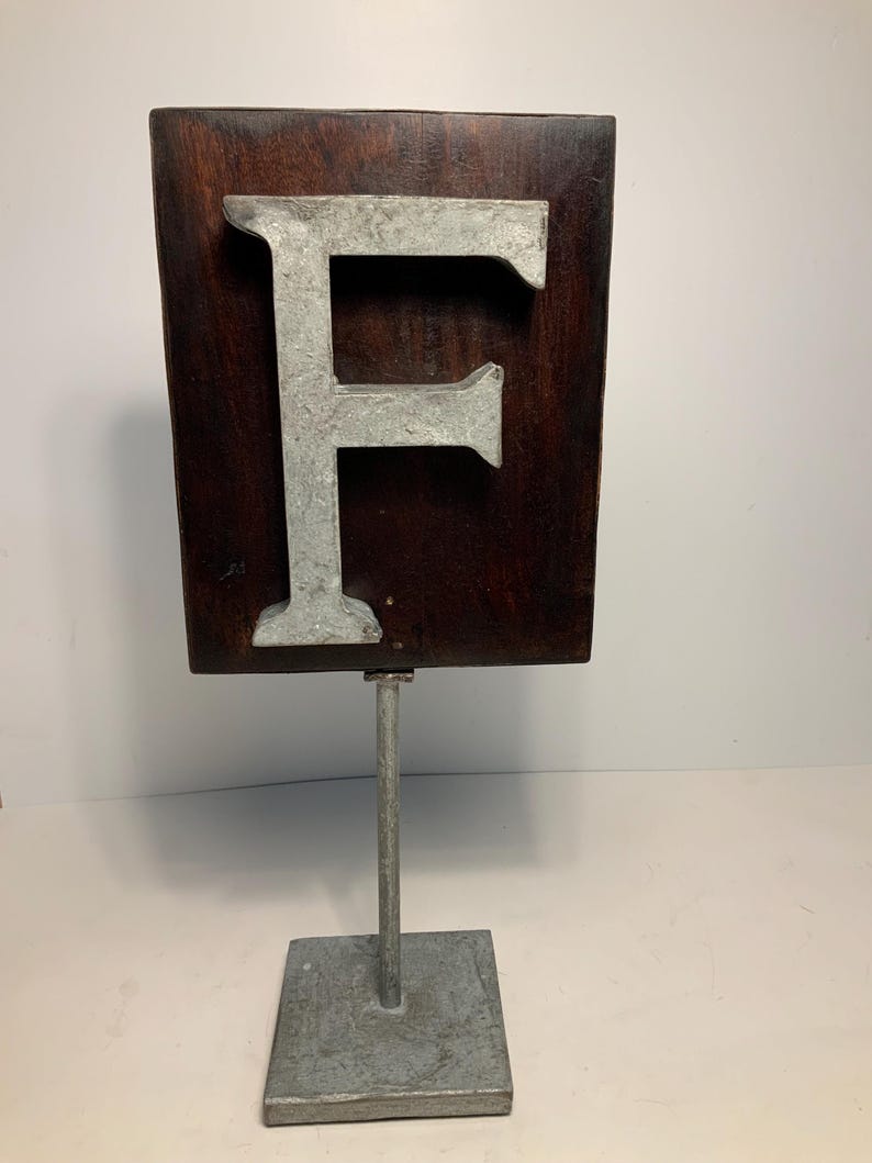 May include: A decorative wooden sign featuring the letter "F" in a silver-colored metal. The letter is mounted on a dark brown rectangular wooden plaque. The plaque is attached to a metal stand with a square base.