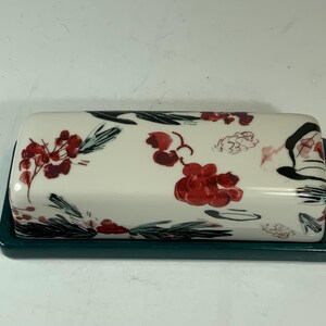 May include: A rectangular ceramic butter dish with a white lid and a dark teal base. The lid is decorated with red berry clusters, green leaves, and black accents. The dish is designed for serving butter.