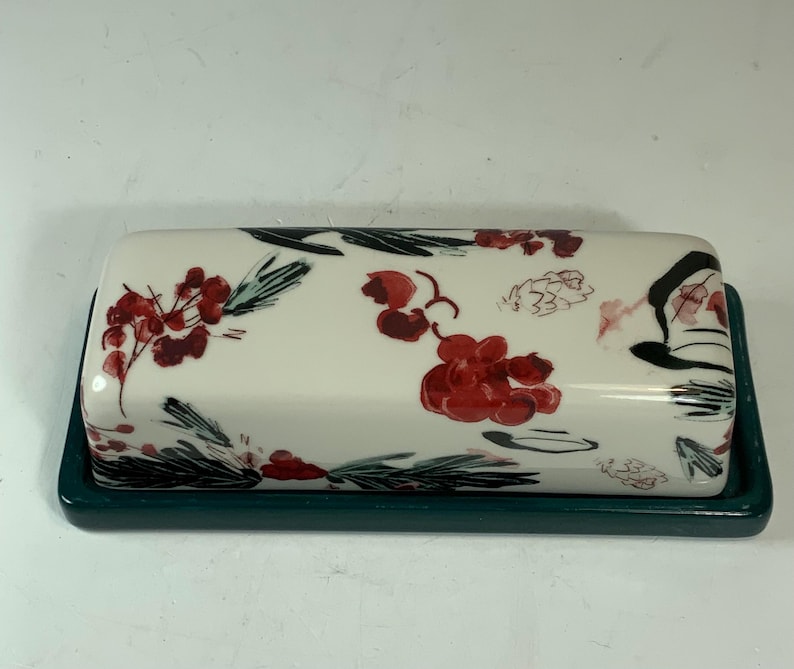 May include: A rectangular ceramic butter dish with a white lid and a dark teal base. The lid is decorated with red berry clusters, green leaves, and black accents. The dish is designed for serving butter.