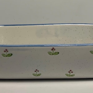 May include: Rectangular ceramic baking dish with a speckled white finish, blue trim, and small floral designs. The dish has handles on each end and is suitable for baking casseroles and other dishes.