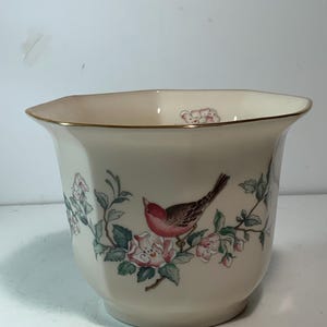 May include: Cream-colored ceramic planter with a gold rim and a hexagonal shape. The planter features a painted design of a bird perched on a flowering branch with pink roses and green leaves. The overall aesthetic is vintage and decorative.