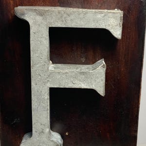 May include: A large, metallic letter "F" is mounted on a dark brown, rectangular wooden plaque. The letter has a weathered, industrial appearance. The plaque is mounted on a metal rod, suggesting a sign or decorative piece.