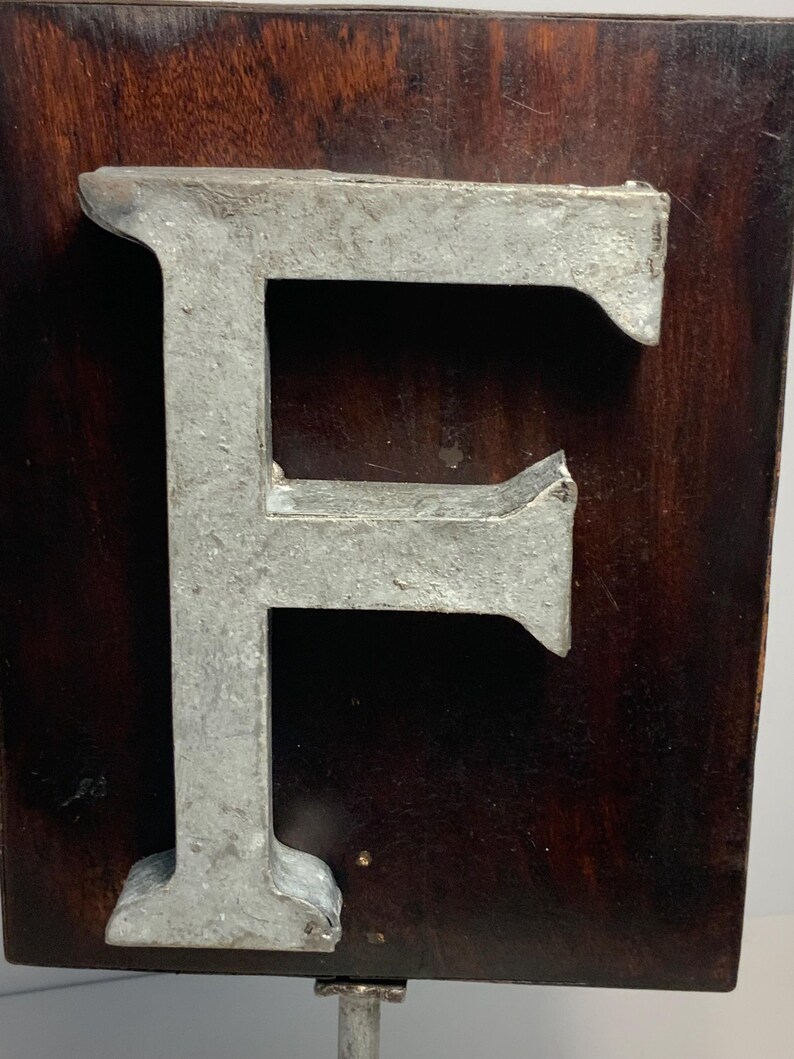 May include: A large, metallic letter "F" is mounted on a dark brown, rectangular wooden plaque. The letter has a weathered, industrial appearance. The plaque is mounted on a metal rod, suggesting a sign or decorative piece.