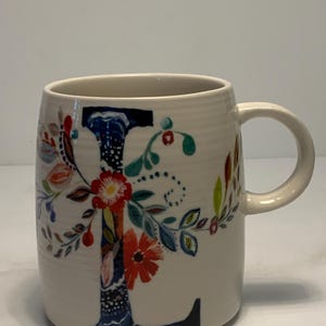 Puede incluir: Taza de cer&aacute;mica color crema con un gran dise&ntilde;o floral de la letra "L". La letra est&aacute; adornada con flores y hojas coloridas en rojo, azul y verde. La taza tiene forma redondeada y un asa curva.