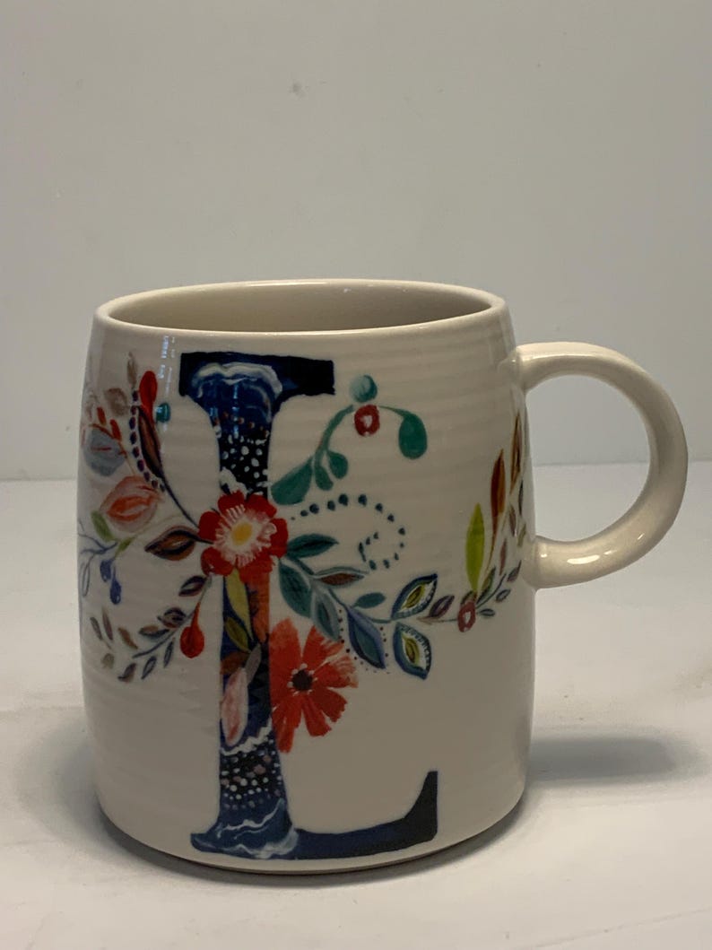 Puede incluir: Taza de cer&aacute;mica color crema con un gran dise&ntilde;o floral de la letra "L". La letra est&aacute; adornada con flores y hojas coloridas en rojo, azul y verde. La taza tiene forma redondeada y un asa curva.