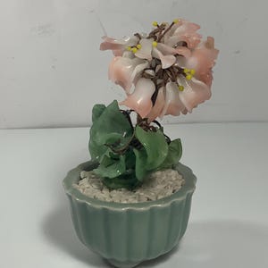 Pode incluir: Uma pequena planta de jade com flores cor-de-rosa num vaso de cerâmica verde. A planta é feita de jade e as flores são feitas de vidro rosa.