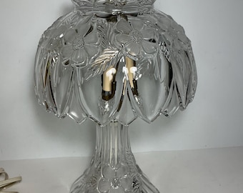 May include: A clear glass table lamp featuring a dome-shaped shade with embossed floral patterns and scalloped edges. The base is also made of clear glass with matching floral details. A silver-toned metal finial sits at the top.