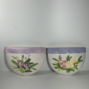 May include: Two white ceramic wall planters with a light purple rim. The planter on the left has a purple and green floral design. The planter on the right has a pink and yellow floral design.