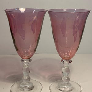 May include: A pair of pink-tinted wine glasses. Each glass features a clear, decorative stem and a flared bowl. The base is a clear, circular disc. The glasses are likely made of glass.
