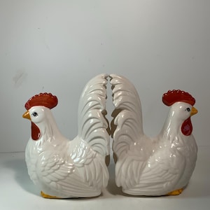 May include: Two white ceramic rooster figurines with red combs and wattles, yellow beaks, and yellow feet. The roosters have detailed feather textures and are positioned facing each other.