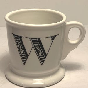 May include: A white ceramic mug featuring a large black letter "W" with floral detailing. The mug has a handle and a wide base. The design is simple and elegant.