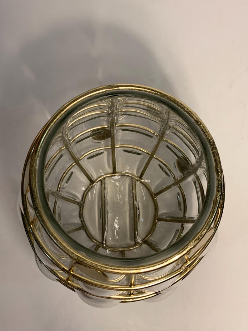 Op de afbeelding: Transparante glazen kom met een goudkleurig metalen kooi-ontwerp. De kom heeft een afgeronde vorm met een decoratieve metalen rand. Het kooi-ontwerp voegt een vintage of antieke esthetiek toe.