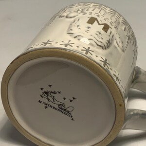 May include: White ceramic mug with a textured design, a gold-colored rim, and a handle. The mug features a gold letter "M" and snowflake accents. The bottom of the mug has the text "by ANTHROPOLOGIE" and a graphic of a reindeer.
