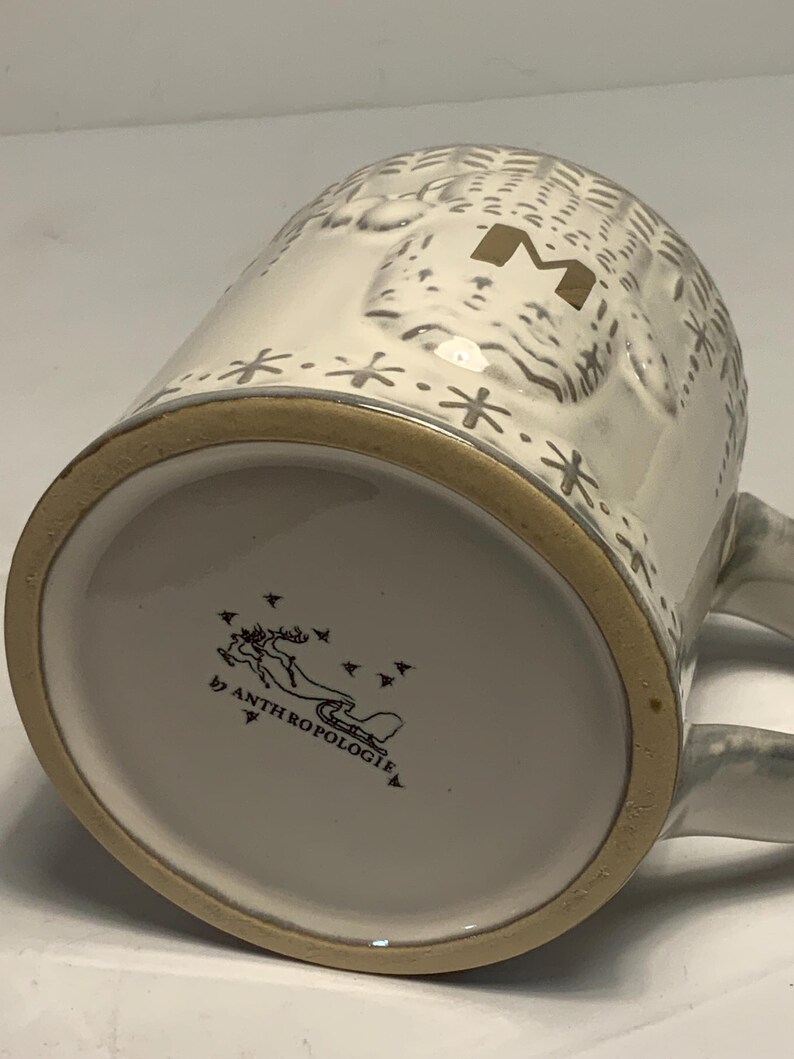 May include: White ceramic mug with a textured design, a gold-colored rim, and a handle. The mug features a gold letter "M" and snowflake accents. The bottom of the mug has the text "by ANTHROPOLOGIE" and a graphic of a reindeer.