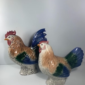 May include: Two ceramic rooster figurines. The roosters have brown, blue, green, and white coloring. The roosters have red combs and yellow beaks. The figurines are on a white surface.