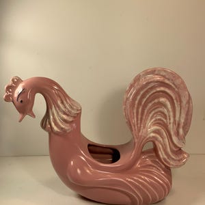 May include: A pink ceramic rooster-shaped planter with a curved neck and a large, fanned tail. The rooster has a small, detailed head with a beak and a crest. The tail features white and pink swirled details.