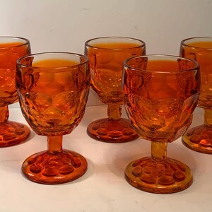 Può includere: Un set di cinque calici in vetro color ambra caratterizzati da un motivo a nido d'ape testurizzato sulla coppa e una base circolare. Ogni bicchiere presenta uno stelo corto e una coppa arrotondata con un motivo geometrico ripetuto.