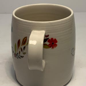 Puede incluir: Taza de cer&aacute;mica color crema con dise&ntilde;o floral. La taza tiene un asa blanca y est&aacute; adornada con patrones florales rojos, marrones y verdes. La taza tiene forma cil&iacute;ndrica y una superficie texturizada.