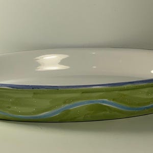May include: Oval ceramic baking dish with a white interior and a green exterior. The green exterior has a blue wavy design and a blue rim. The dish is approximately 30 cm long and 20 cm wide.