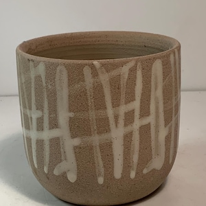 May include: A handcrafted, beige ceramic bowl with a rounded shape. The bowl features a textured surface and is decorated with vertical and horizontal white glaze lines, creating a grid-like pattern. The interior is a lighter beige.