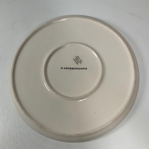 May include: Off-white ceramic plate with a slightly raised edge. The underside features the text "by ANTHROPOLOGIE" in black, along with other text and a circular design. The plate is on a white surface.