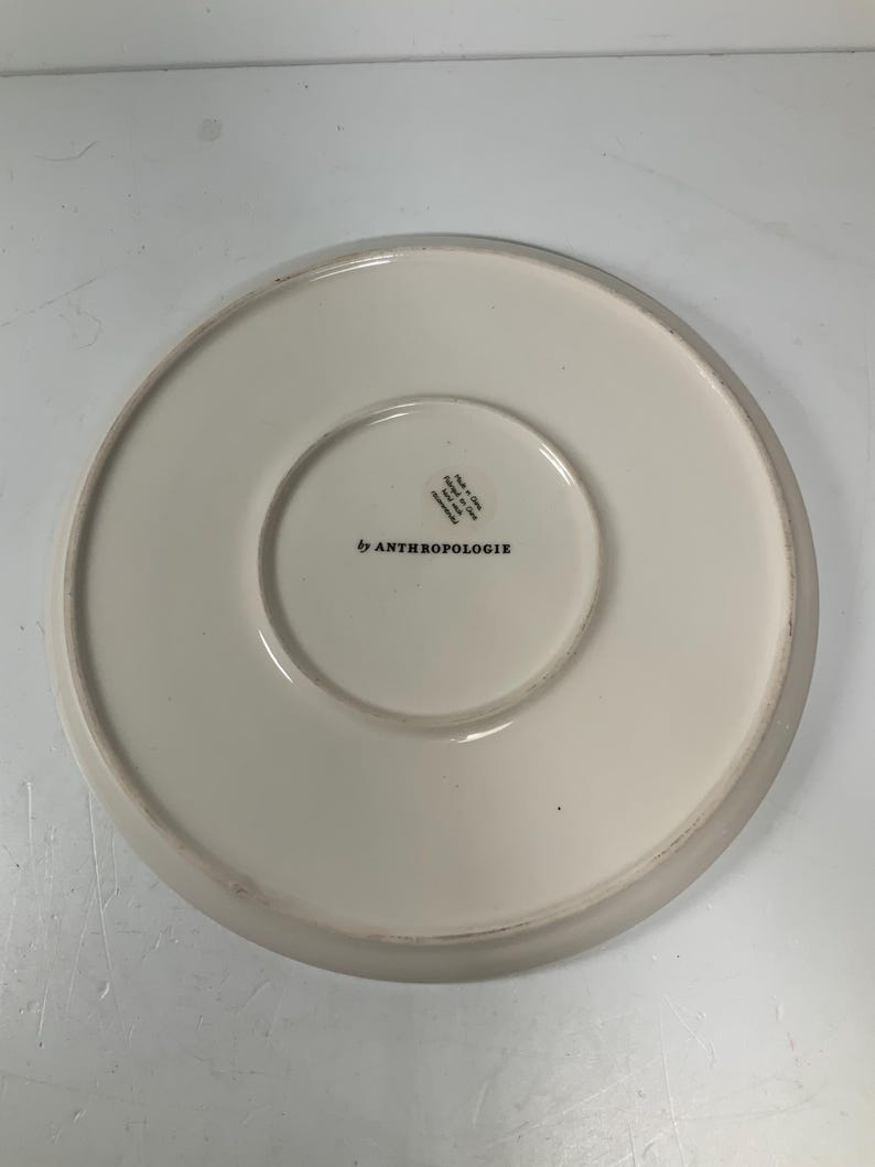 May include: Off-white ceramic plate with a slightly raised edge. The underside features the text "by ANTHROPOLOGIE" in black, along with other text and a circular design. The plate is on a white surface.