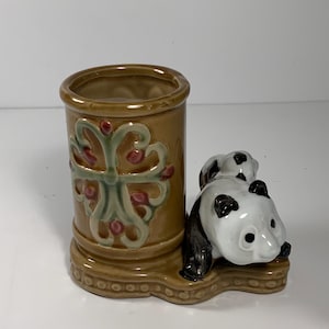 May include: A ceramic panda bear figurine with a brown cylindrical container. The container has a decorative floral design in green, pink, and white. The panda bears are black and white.