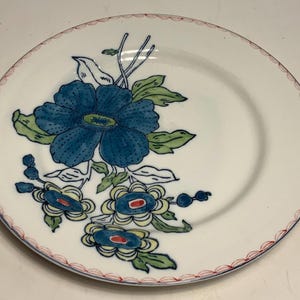 May include: White ceramic plate with a blue floral design. The plate features a large blue flower with green leaves and smaller blue and yellow flowers. The rim is decorated with a red and white scalloped pattern.