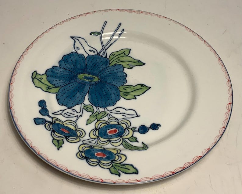 May include: White ceramic plate with a blue floral design. The plate features a large blue flower with green leaves and smaller blue and yellow flowers. The rim is decorated with a red and white scalloped pattern.