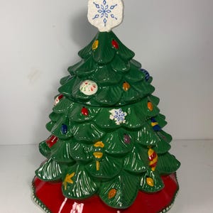 May include: A ceramic Christmas tree cookie jar. The tree is green with colorful lights and ornaments. It has a red base and a white snowflake on top. The jar is designed to resemble a traditional Christmas tree.