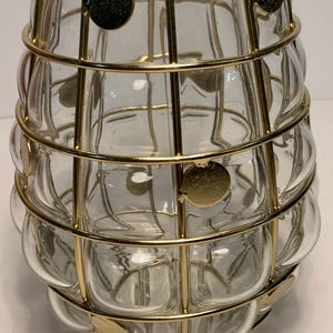 Op de afbeelding: Een heldere glazen vaas met een bubbelachtige textuur, omhuld door een gouden metalen kooi. De kooi heeft cirkelvormige gouden accenten. De vaas heeft een uniek, decoratief ontwerp, geschikt voor het tentoonstellen van bloemen of als een op zichzelf staand kunstwerk.
